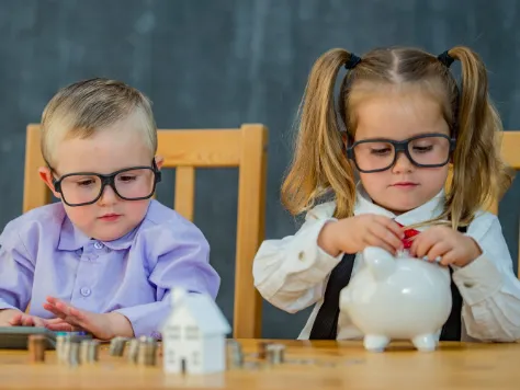 Zwei Kinder in Erwachsenenkleidung sitzen am Tisch; das Mädchen steckt Münzen in ein Sparschwein, während der Junge einen Taschenrechner hält und Münzstapel vor ihnen liegen.