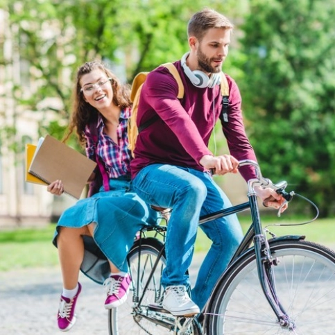 Zwei junge Menschen fahren auf einem Fahrrad durch einen Park, halten Ordner und Hefte, grüne Bäume im Hintergrund.