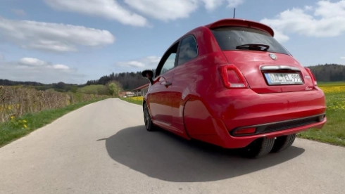 Roter Fiat 500 auf ländlicher Straße, grüne Felder und Wald im Hintergrund, sonniger Tag