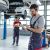 In einer KFZ-Werkstatt stehen stehen Autos auf einer Hebebühne. Zwei Kfz-Mechaniker arbeiten in der Werkstatt,  einer der Kfz-Mechaniker tippt etwas in sein Tablet .