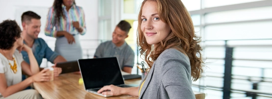 Frau mit Laptop arbeitet am Tisch, während Kollegen im Hintergrund in modernem Büro zusammenarbeiten