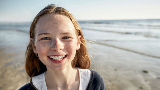 Lächelndes Mädchen mit Sommersprossen am Strand, Meer im Hintergrund, sonniger Tag