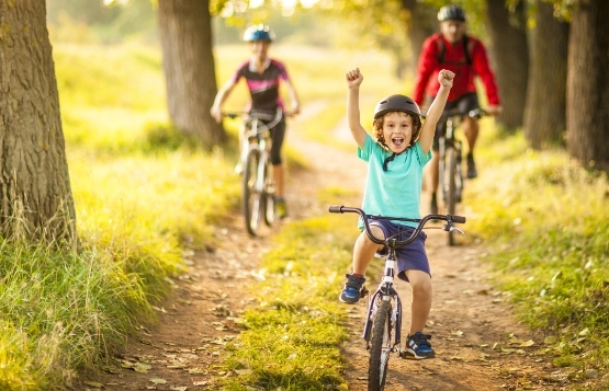 Kind auf Fahrrad fährt freudig auf Waldweg, Familie mit Kindern folgt auf Rädern durch sonnigen Wald.