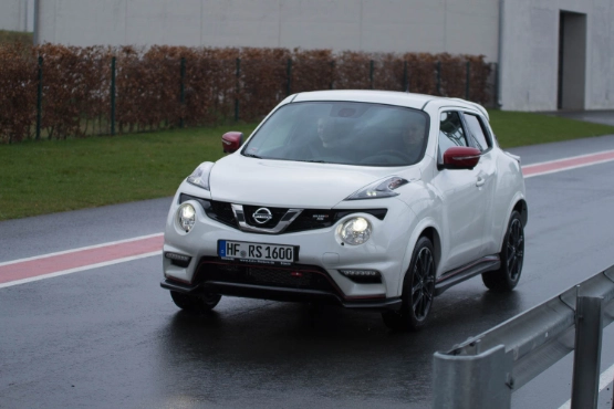 Weißer Nissan Juke fährt auf nasser Straße, Grünfläche und Gebäude im Hintergrund