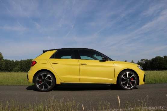 Gelber Audi A1 Sportwagen in Seitenansicht auf leerer Straße, grüne Landschaft im Hintergrund, blauer Himmel