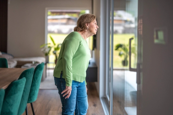 Seniorin im gruenen Pullover die durch das Fenster im Wohnzimmer schaut