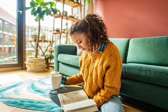 Frau in gelbem Pullover sitzt auf grünem Sofa, arbeitet am Laptop mit Kaffeetasse in modernem Wohnzimmer