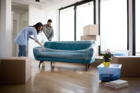 Zwei Personen positionieren ein türkises Sofa in hellem Wohnzimmer mit großen Fenstern und Umzugskartons