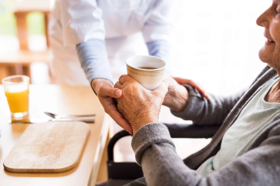 Pflegekraft reicht vorsichtig einer sitzenden aelteren Frau einen Kaffeebecher