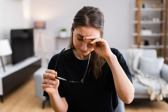 Frau mit Brille in schwarzem Shirt sitzt am Schreibtisch und reibt sich erschöpft die Augen, Büroumgebung im Hintergrund