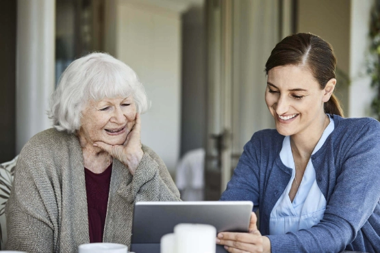 Pflegerin sitzt mit Seniorin am Tisch und schauen gemeinsam auf ein Tablet