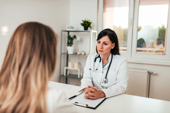 Ärztin im weißen Kittel mit Stethoskop führt Patientengespräch am Schreibtisch in moderner Arztpraxis.