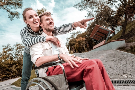 Frau laechelt und umarmt ihren Freund der im Rollstuhl sitzt bei einem Ausflug