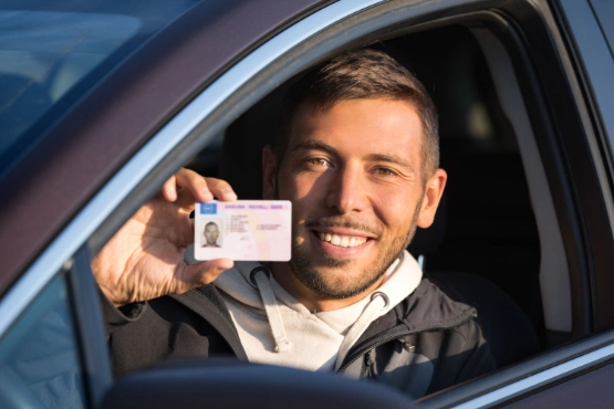 Mann im Auto hält Führerschein oder Dokument in die Kamera und lächelt.