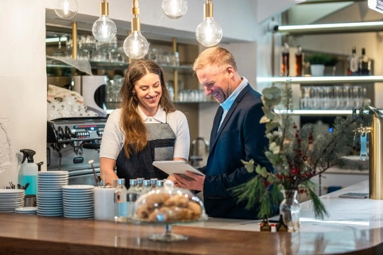 Barista mit Tablet und Kunde in moderner Kaffeebar mit Regalen und Pflanzen