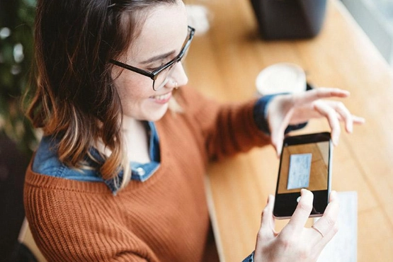 Frau mit Brille hält Smartphone in den Händen und fotografiert damit auf Holztisch.