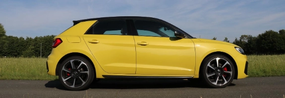 Gelber Audi A1 Sportwagen in Seitenansicht auf leerer Straße, grüne Landschaft im Hintergrund, blauer Himmel