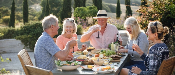 Gruppe älterer Menschen sitzt an Tisch mit Speisen im mediterranen Garten, genießt gemeinsames Essen und Gespräche