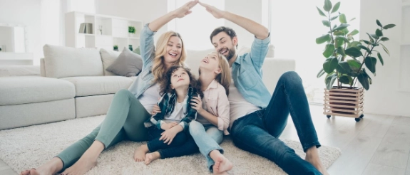 Familie sitzt auf Teppich im Wohnzimmer, bildet Dach mit Händen über Köpfen, modernes helles Interieur mit Sofa und Pflanze