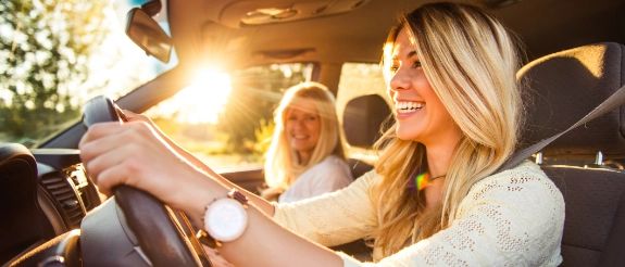 Blonde Frau steuert lachend ein Cabrio-Auto, während Freunde neben ihr sitzen und die Sonne scheint