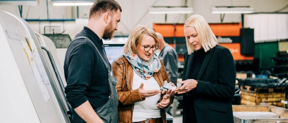 Drei Personen in einer Fabrikhalle besprechen sich vor technischen Zeichnungen und schauen auf ein Tablet.