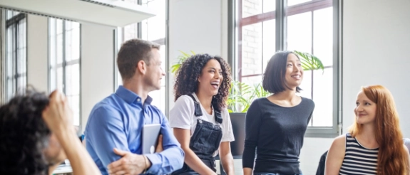 Diverse Gruppe von fünf Personen steht in hellem Büro zusammen und schaut nach rechts, helles Tageslicht von großen Fenstern.