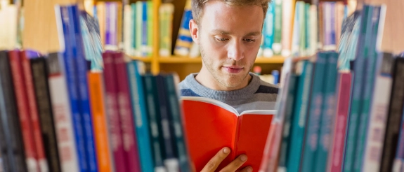 Junge mit rotem Buch in Bibliothek zwischen bunten Bücherregalen lesend