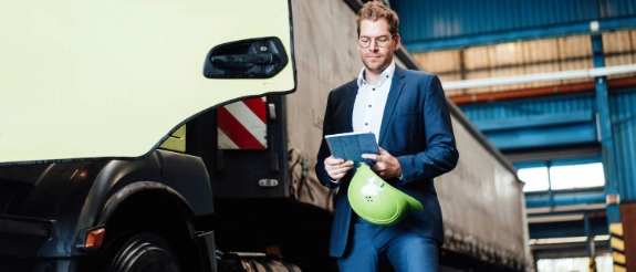 Mann in blauem Anzug mit Tablet und gelbem Helm vor Lastwagen in Lagerhalle