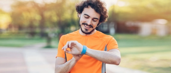 Mann in orangem T-Shirt blickt auf blaues Fitness-Armband, lächelt, outdoor grüner Hintergrund