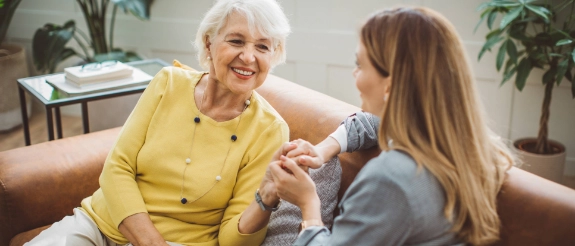 Ältere Frau sitzt lächelnd auf dem Sofa und hält die Hände einer jüngeren Frau die sie fürsorglich anblickt – Symbolbild für pflegende Angehörige familiäre Unterstützung und Pflege zuhause