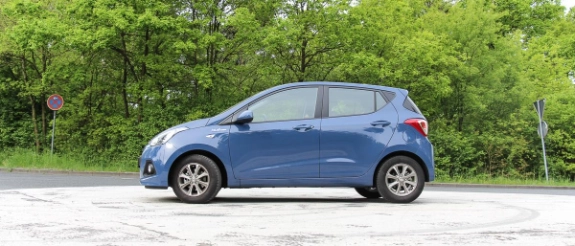 Blauer Kleinwagen steht auf grauem Parkplatz vor grünem Wald und Bäumen