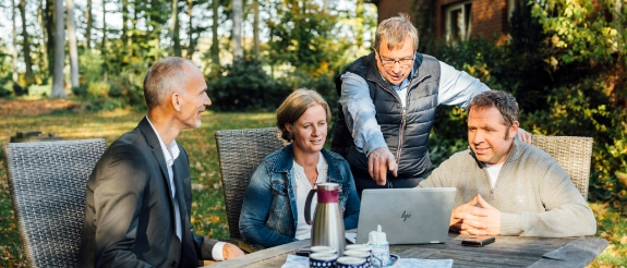 Vier Geschäftsleute besprechen sich outdoor an Tisch mit Laptop, Thermosflasche und Unterlagen im grünen Garten.