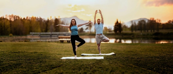Ein Paar praktiziert Yoga im Freien. Im Hintergrund ist ein See, eine Bank und schöne Landschaft zu sehen.