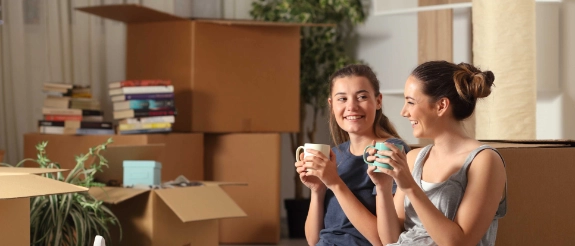 Zwei Frauen sitzen auf dem Boden, trinken Kaffee und feiern ihren Umzug zwischen Umzugskartons in ihrer neuen Wohnung.