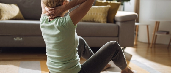 Frau macht Sit-ups auf Fitnessmatte im Wohnzimmer, Sofa und Pflanzen im Hintergrund