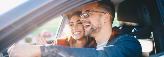 Junges Paar sitzt lächelnd im Auto, Mann am Steuer, Frau schaut aus dem Fenster, sonniges Wetter