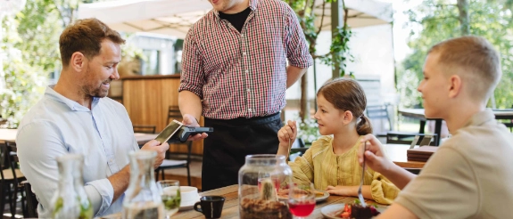 Familie sitzt am Esstisch in moderner Küche, Vater steht und serviert Essen, Kinder und Mutter lachen zusammen