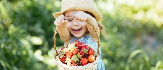 Kleines Mädchen mit Strohhut hält Korb voller frischer Erdbeeren und Obst im grünen Garten