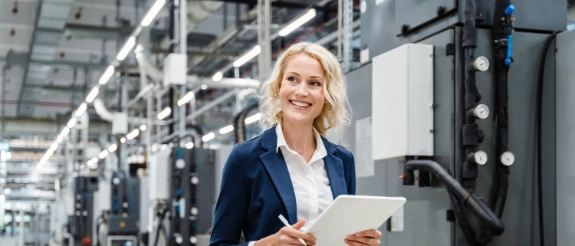 Blonde Frau in Businessanzug mit Tablet in moderner Industriefabrik, lächelnd zwischen Maschinen.
