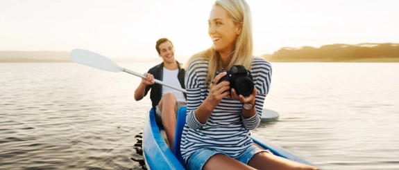 Frau mit Kamera in Kajak lächelt, Mann mit Paddel im Hintergrund, Wasser und Uferlandschaft bei Sonnenuntergang