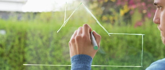 Person zeichnet Hausumriss auf Fenster mit grünem Garten im Hintergrund