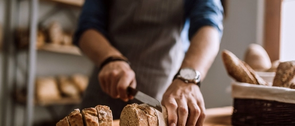 Handwerker in Schürze formt Brot auf Holztisch, Bäckerei im Hintergrund sichtbar