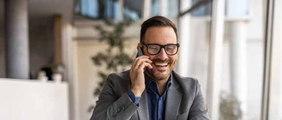 Mann im grauen Anzug und Brille telefoniert lächelnd in modernem Büro mit Pflanze im Hintergrund.