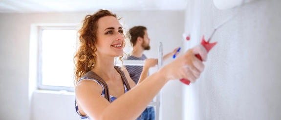 Frau mit roten Haaren streicht weiße Wand mit Farbrolle, Mann im Hintergrund, helles Wohnzimmer während Renovierung