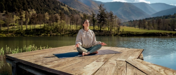 Junge Frau in Trainingsanzug sitzt auf einem Steg am See und praktiziert Yoga.