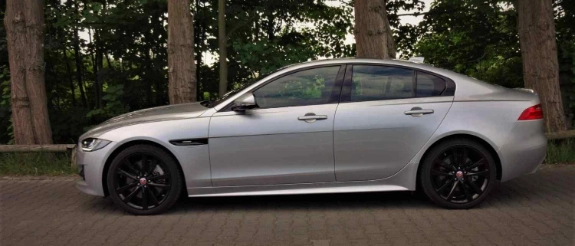 Silberner BMW M340i Limousine mit schwarzen Felgen steht seitlich auf Parkplatz vor Bäumen