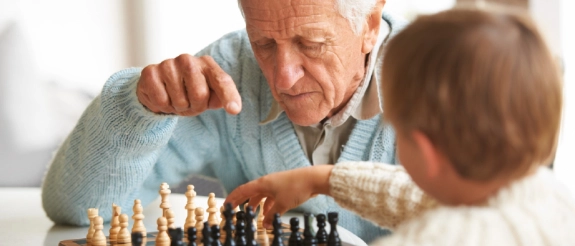 Großvater und Enkelin spielen Schach zusammen an einem Holztisch in heller Wohnumgebung.