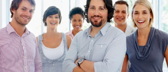 Professionelles Team von sechs Personen steht lächelnd zusammen in moderner, heller Büroumgebung mit großen Fenstern