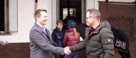 Zwei Männer schütteln sich die Hand vor einem Gebäude, im Hintergrund weitere Personen in Winterkleidung.