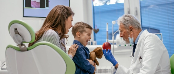 Zahnarzt untersucht lachendes Kind im Behandlungszimmer, Mutter sitzt daneben, moderne Zahnarztpraxis mit Monitoren